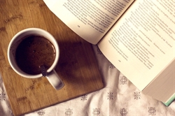 [ai] A white coffee cup filled with dark coffee sits on a wooden cutting board. An open book with printed text is partially visible beside the cup on a patterned tablecloth.
